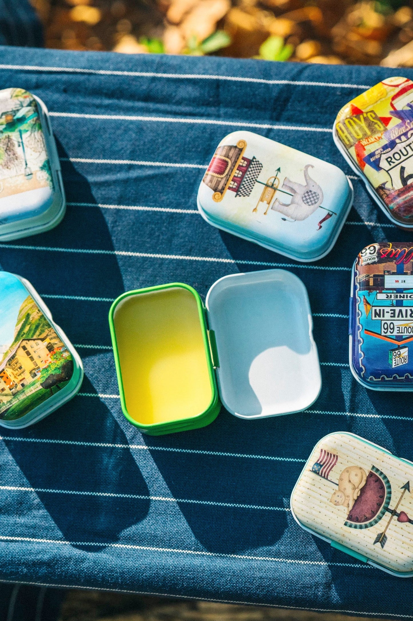 Soothing salve tins on a pin-stripe blue and white tea towel. One tin is open, showing the creamy salve contents. The other tins display vintage designs.