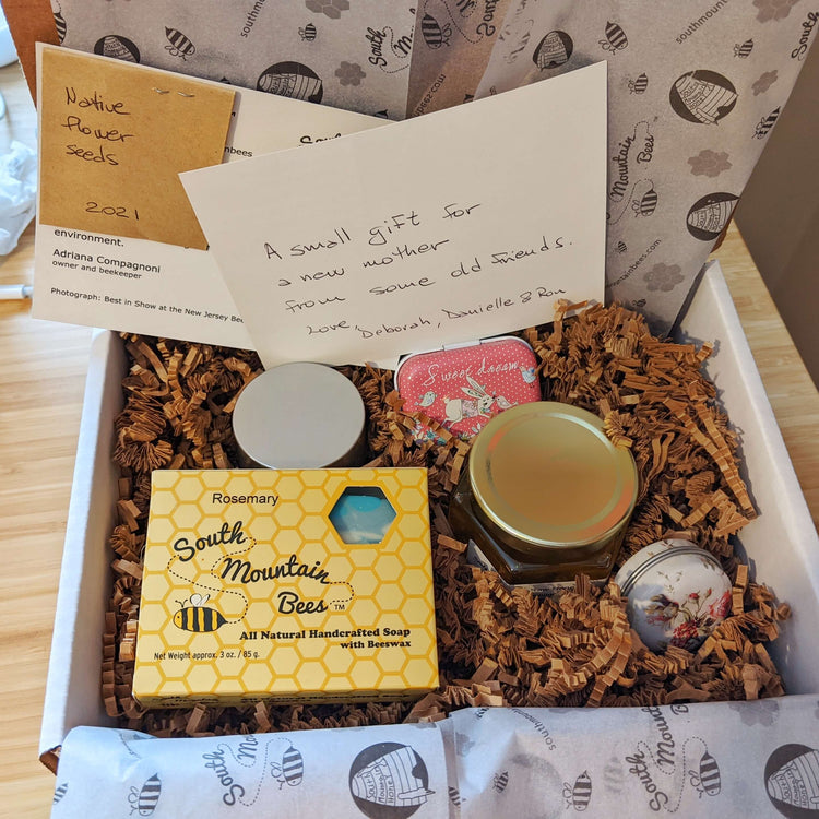 Gift box of natural cosmetics: honey soap, beeswax lip balm, honey scrub with bamboo spoon, soothing salve, moisturizer with Shea butter and Vitamin E, free seeds, and a handwritten message.