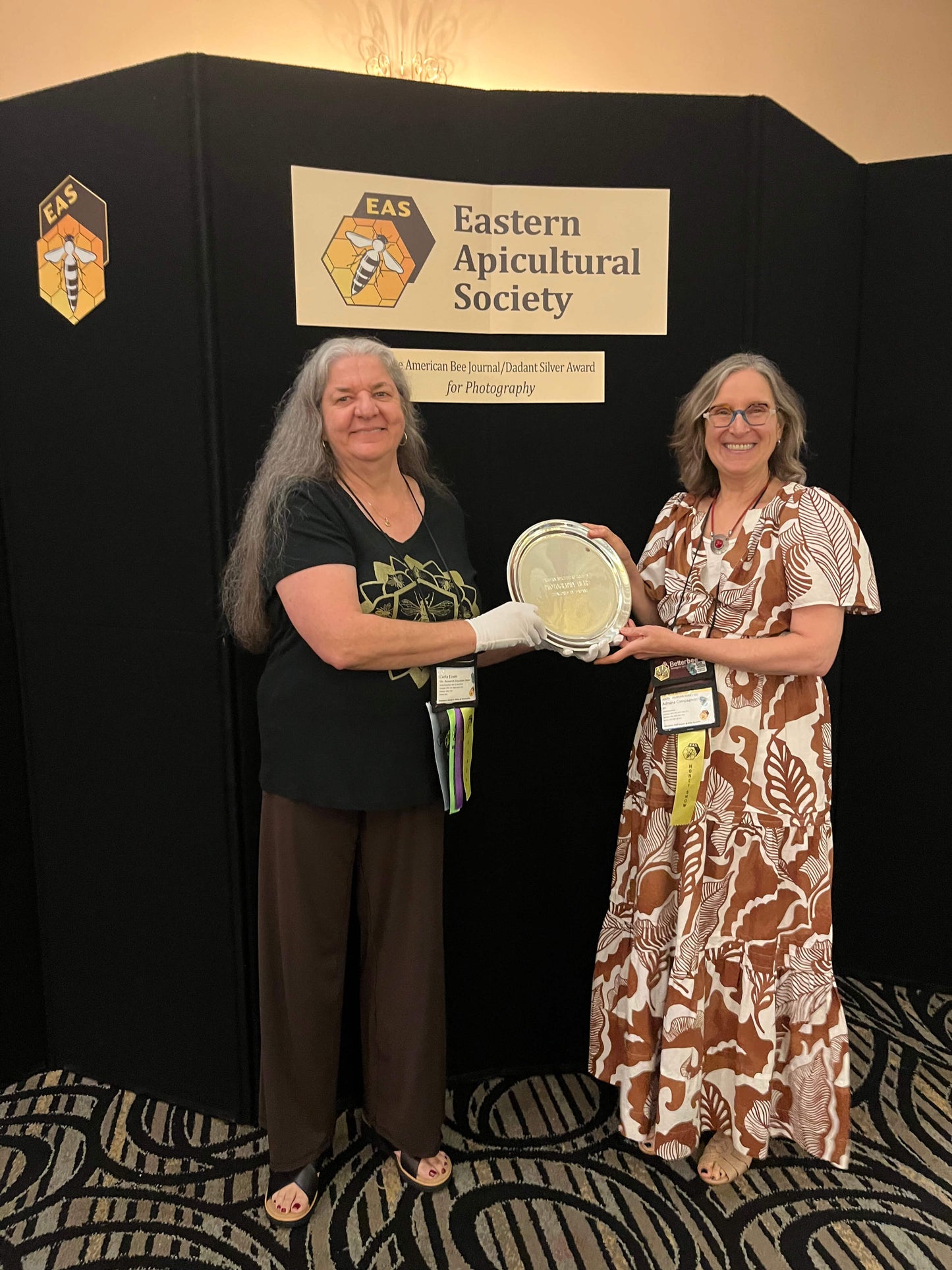 Adriana and the conference chair holding the silver award plaque in front of an Eastern Apicultural Society backdrop
