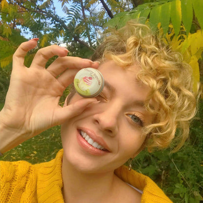 Beautiful woman holding a lovely tin of our award winning lip balm in front of her closed right eye.