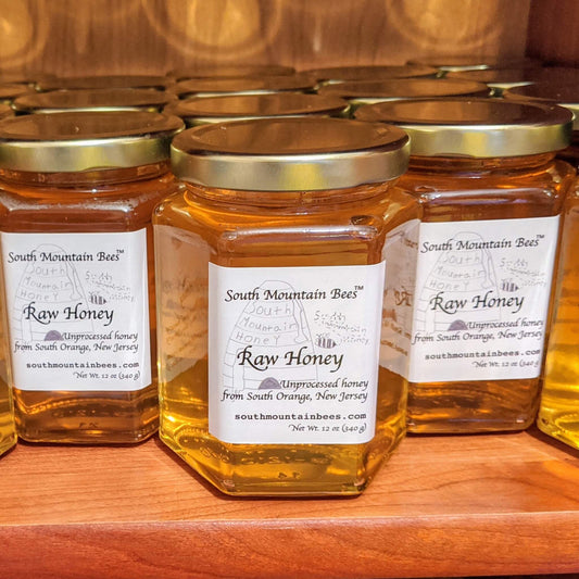 12 ounces of delicious golden honey in a glass hexagon jar rests on a wooden shelf.