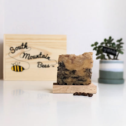 A bar of espresso honey soap has freckles of coffee on a honey and beeswax soap base. The soap sits on a beech-wood soap dish, with a few espresso beans on the foreground, and a branded wooden box and a potted succulent in the background.