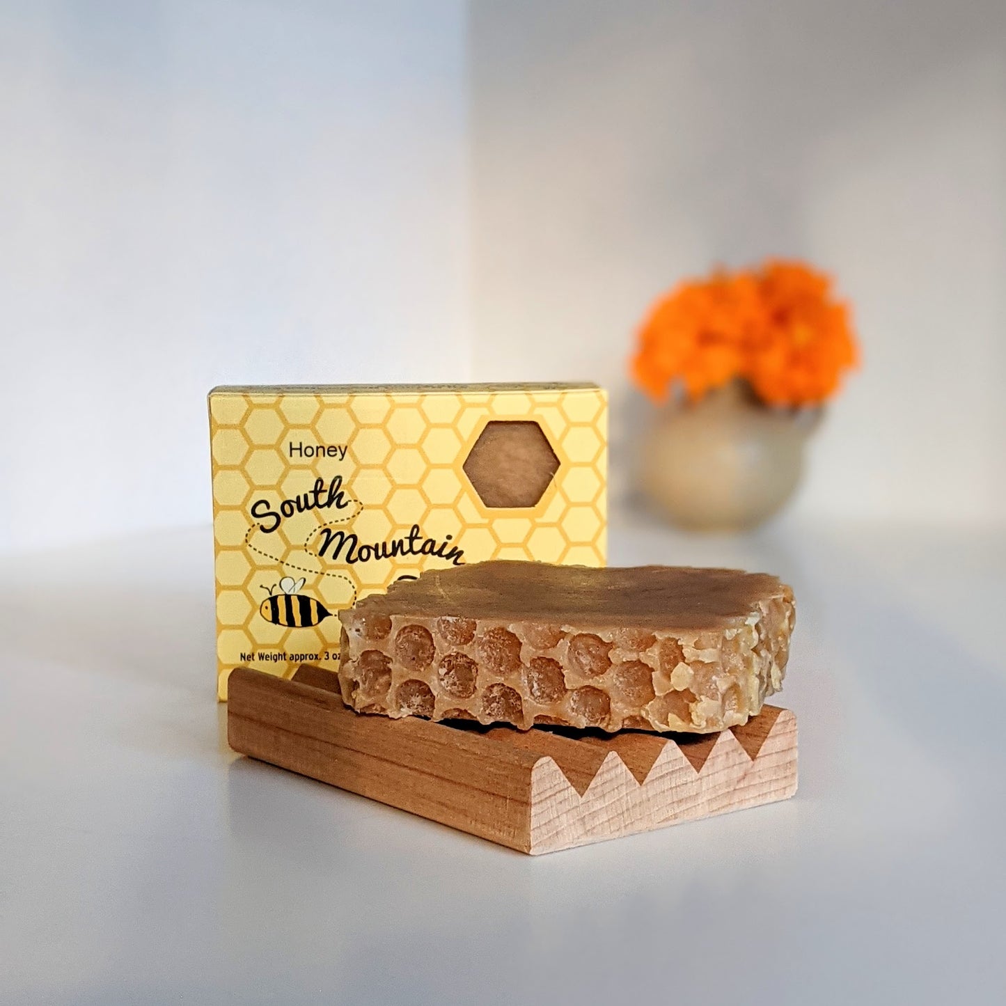 A honey soap on a wooden soap dish next to a honey soap box with the signature hexagon window to peek and smell the soap while protecting it. Marigolds fade in the background.