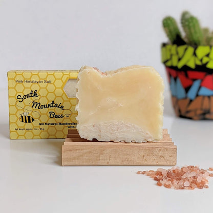 A bar of natural pink Himalayan soap on a birch wood soap dish. A soap in its box right behind it. A handful of pink salts in the foreground and a potted cactus in the background.