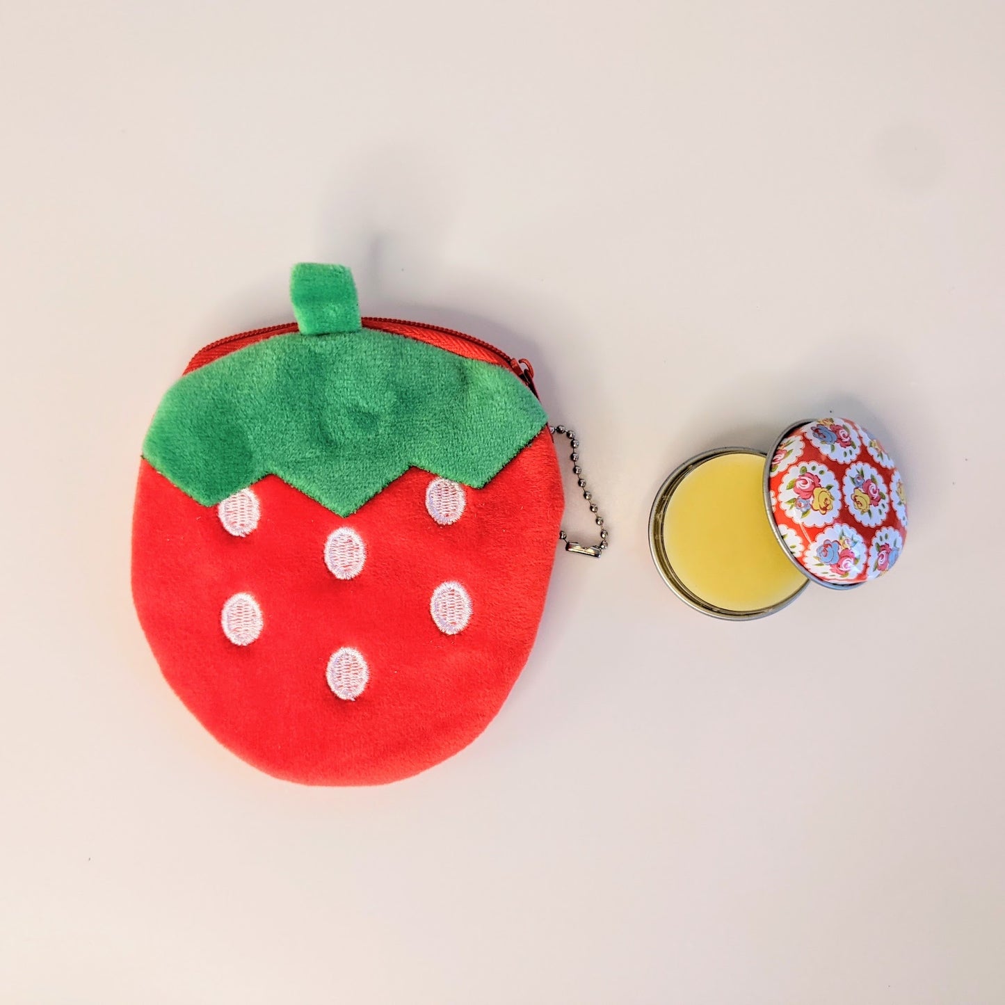 Soft plush bright red and green strawberry shaped coin purse next to open lip balm tin.