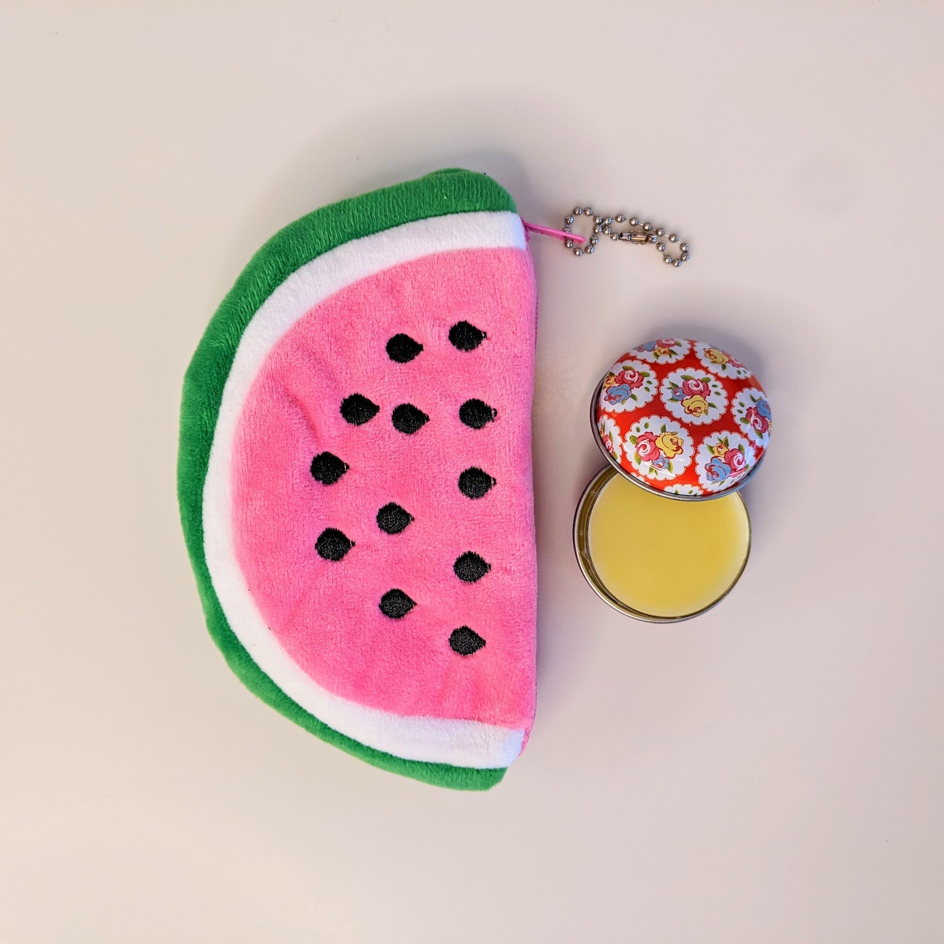 A soft plush coin purse in the shape of a watermelon slice next to an open tin of beeswax lip balm.