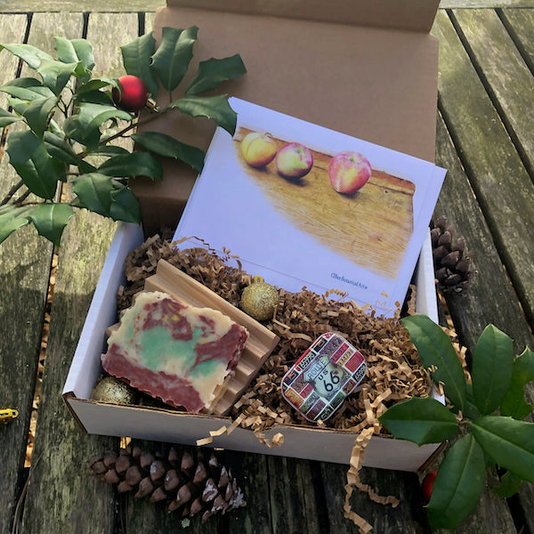 A gift box with a honey soap, a wooden soap dish, a salve, and a set of 4 cards: three apples on wood table.