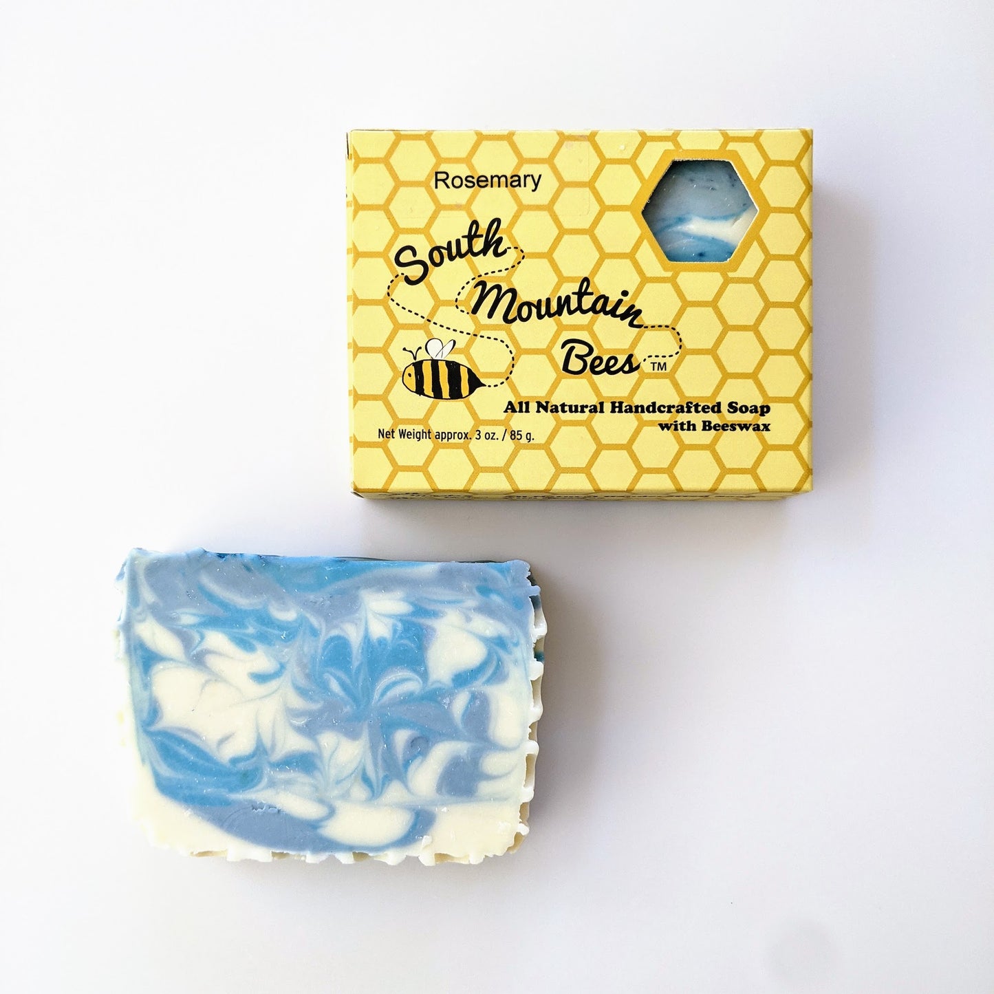 Rosemary Dream is a honey soap with blue and grey swirls. It sits next to a soap box where you can see the soap through a hexagonal window.