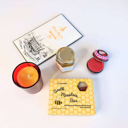 These are the contents of the Sweet Candle Light  gift box. It is packed with delightful products: a honey soap, a 2oz raw honey jar from our bees, a 100% beeswax votive candle for over 12 hours of burning time in a red glass candle holder, a cherry lip gloss in a metal tin, and a craft chocolate bar.